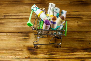 Shopping cart with money from different countries: euros, us dollars, ukrainian hryvnia, russian rubles, egyptian pounds on wooden background