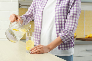 Wall Mural - Young woman pouring lemon water into glass from jug in kitchen, closeup