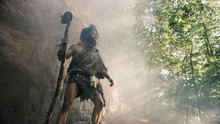 Primeval Caveman Wearing Animal Skin Holds Stone Hammer Stands Near Cave And Looks Around Prehistoric Landscape, Ready To Hunt Animal Prey. Neanderthal Going Hunting Into Jungle. Low Angle Shot
