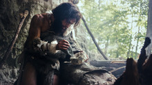Primeval Caveman Wearing Animal Skin Hits Rock With Sharp Stone And Makes First Primitive Tool For Hunting Animal Prey Or To Handle Hides. Neanderthal Using Handax. Dawn Of Human Civilization