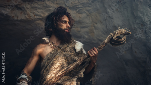 Primeval Caveman Wearing Animal Skin Holds Stone Hammer Stands Near Cave and Looks Around Prehistoric Landscape, Ready to Hunt Animal Prey. Neanderthal Going Hunting into Jungle. Low Angle Shot