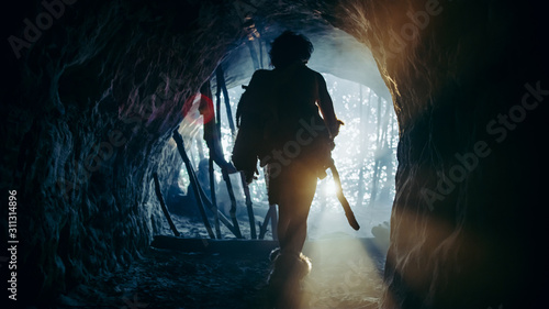 Primeval Caveman Wearing Animal Skin and Fur Holds Stone Tipped Spear Comes out of His Cave into Prehistoric Forest Ready to Hunt. Neanderthal Going Hunting into the Jungle. Shot with Cold Filter.
