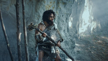 Primeval Caveman Wearing Animal Skin Holds Stone Tipped Hammer Comes Out Of The Cave And Looks Around Prehistoric Forest, Ready To Hunt Animal Prey. Neanderthal Going Hunting Into The Jungle.