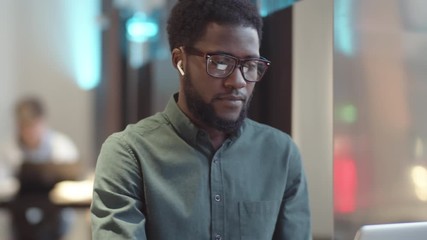 Tilting down shot of focused African American man with beard sitting in restaurant and working on laptop, and reflection of computer screen on his glasses