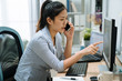 © PR Image Factory - frowning asian japanese woman employee working on laptop computer at office while talking on mobile phone in light workplace. serious young girl discussing project on cellphone online point monitor