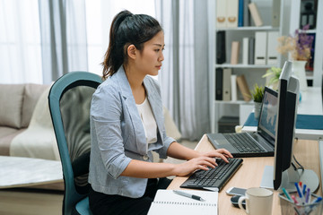 side view charming asian korean girl working at home on computer using her laptop also. female self employed freelancer lifestyle concept. beautiful elegant woman typing on keyboard in living room.