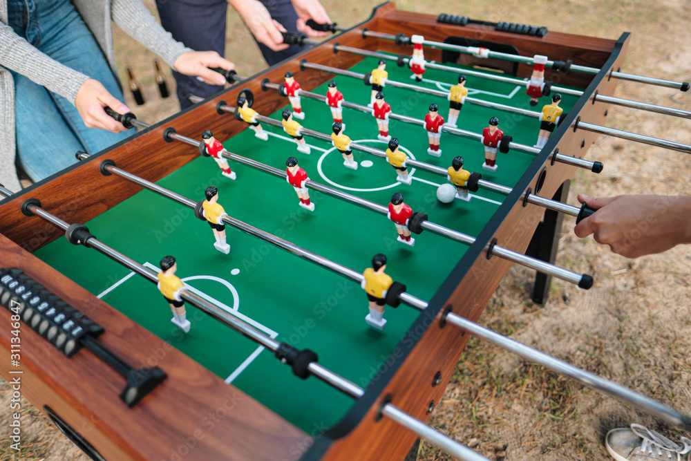 Friends play table football or kicker outdoors. Players and fans rejoice in the victory. - obrazy, fototapety, plakaty 