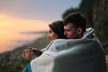 A couple in love sits on the riverbank at sunset. They cuddle, roll into a plaid and drink tea from a metal hiking mug.