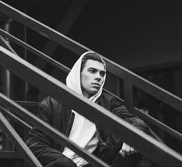 Wall Mural - Beautiful stylish man in black jacket, white hoodie sitting on the stairs