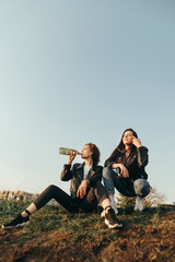 Wall Mural - Two stylish girlfriends sitting on a hill on sky background drinking wine at sunset.Two girls on a walk drinking wine and relaxing,sitting on the ground.Fashionable photo of girls drinking wine