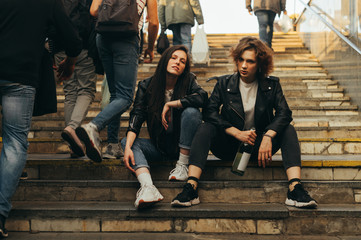 Wall Mural - Portrait of two free girls in stylish clothes sitting on the stairs with a bottle of wine in hand in the middle of a crowd of people. Stylish models posing at the exit of subway,sitting on the stairs