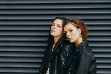 Wall Mural - Portrait of two stylish beautiful girls in casual clothes on a black wall background, looking in camera with a serious face. Two models in leather jackets posing for the camera, dark wall background