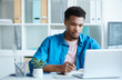 © AnnaStills - Serious African young man in casual clothing sitting at the table looking at monitor of laptop and making notes at office