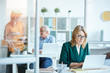 © AnnaStills - Serious mature woman in eyeglasses typing on laptop computer at desk at office with business people in the background