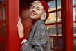 Fashionable happy smiling blonde woman wearing red beret, checkered coat, white wrist watch, posing in retro street phone booth. Copy, empty space for text