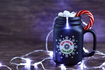 Mug of hot coffee with milk, marshmallow and Lollipop against rustic wooden background with Christmas decorations and lights.