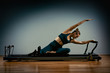 © Georgii - Young girl doing pilates exercises with a reformer bed. Beautiful slim fitness trainer on reformer gray background, low key, art light. Fitness concept