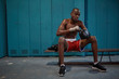 © AnnaStills - Serious African young boxer wearing boxing gloves while sitting on bench in locker room