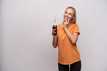 Wall Mural - Indoor photo of cheerful young pretty blonde female in casual clothes holding bottle of soda in raised hand and keeping forefinger on underlip while looking aside with interest