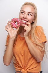 Wall Mural - Pleasant looking young long haired blonde lady in orange t-shirt holding pink donat and smiling happily while keeping forefinger on her underlip, standing against white background