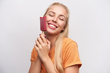 Wall Mural - Pleased good looking pretty young blonde female in casual clothes holding ice-cream on stick and smiling sincerely with closed eyes, standing against white background