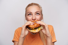 Portrait Of Pleased Young Lovely Blonde Woman With Casual Hairstyle Eating Fresh Hamburger With Great Appetite And Looking Cheerfully Upwards, Posing Over White Background