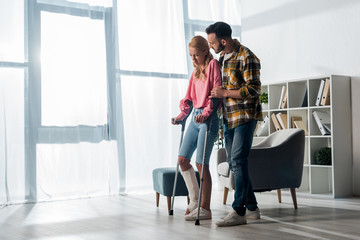 Wall Mural - bearded man looking at injured woman holding crutches at home