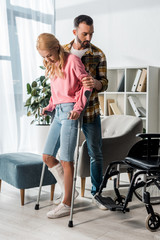 Wall Mural - injured woman holding crutches while standing with man near wheelchair at home