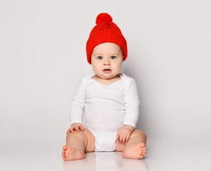 Infant baby boy toddler in white bodysuit and red warm hat with pompon is sitting looking directly at camera on white