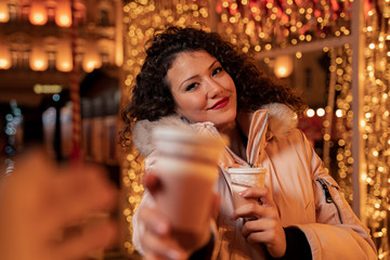 Pretty curly hair girl in the city decorated with Christmas lights holding two cups of coffee to go 