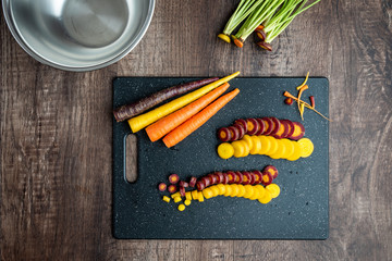 Wall Mural - Sliced rainbow carrots on a black cutting board, stainless-steel bowl, wood table