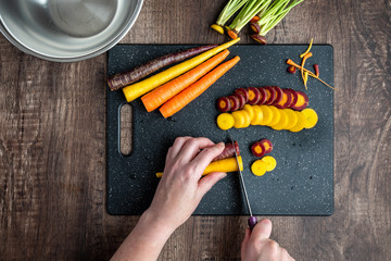 Wall Mural - Woman’s hands cutting rainbow carrots on a black cutting board, stainless-steel bowl, wood table