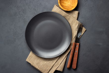 Black Plate And Cutlery Over Napkin On Dark Background. Top View.