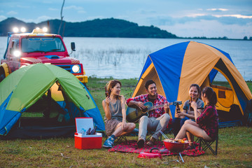 Camping of happy asian young travellers at lake, asian man and women group, relaxing, sing a song and cooking, at sunset.