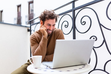 Wall Mural - Happy young man smiling, as he works on his laptop to get all his business done early in the morning with his cup of coffee