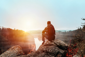 Young man in jacket, pants and backpack sitting on rock looking to river Vltava and valley in sunset.