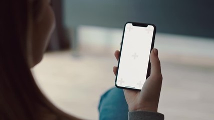 Over shoulder shot of caucasian woman holding a green screen mobile phone or smartphone in her hand. Girl using smartphone in home, surfing the internet, tapping, watching video, blogs, content. Close