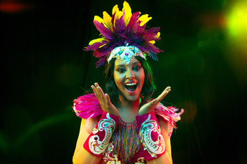 Wall Mural - Beautiful young woman in carnival mask and stylish masquerade costume with feathers in colorful lights and glow on black background. Christmas, New Year, celebration. Festive time, dance, party.