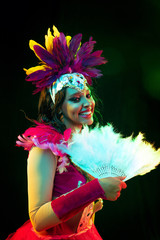 Wall Mural - Beautiful young woman in carnival mask and stylish masquerade costume with feathers fan in colorful lights and glow on black background. Christmas, New Year, celebration. Festive time, dance, party.