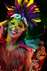 Wall Mural - Beautiful young woman in carnival mask and stylish masquerade costume with feathers in colorful lights and glow on black background. Christmas, New Year, celebration. Festive time, dance, party.