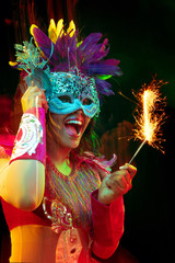 Wall Mural - Beautiful young woman in carnival mask and stylish masquerade costume with feathers and sparklers in colorful lights on black background. Christmas, New Year, celebration. Festive time, dance, party.