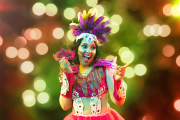 Beautiful young woman in carnival mask and stylish masquerade costume with feathers and sparklers in colorful bokeh on black background. Christmas, New Year, celebration. Festive time, dance, party.