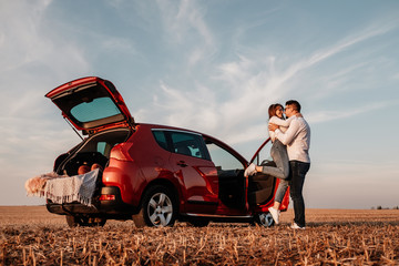 Wall Mural - Young Happy Couple Dressed Alike in White Shirt and Jeans Enjoying Road Trip at Their New Car, Beautiful Sunset on the Field, Vacation and Travel Concept