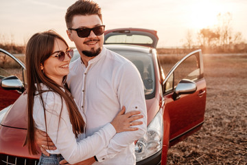 Wall Mural - Young Happy Couple Dressed Alike in White Shirt and Jeans Enjoying Road Trip at Their New Car, Beautiful Sunset on the Field, Vacation and Travel Concept