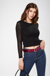 Medium full shot of a brown-haired lady in a black blouse and blue jeans with a red velvet belt with a golden buckle and eyelets. The girl is posing on the gray background. 