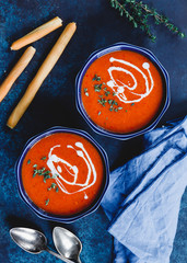 Top view of two portions of red tomato cream soup in bowls with spices on a blue background.