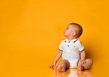 Infant Baby Boy Toddler Is Sitting On The Floor Looking Up At Free Copy Space In Corner Yellow Background