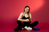 Fototapeta Panele - Young brunette girl in a black top with a headphones and phone sitting in studio on a red cherry background. Near her is a yoga mat and dumbbells