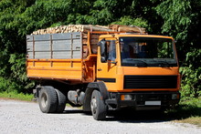 Old Yellow Truck Filled With Firewood Parked On Gravel Parking Lot Next To Public Road Surrounded With Dense Trees On Warm Sunny Summer Day