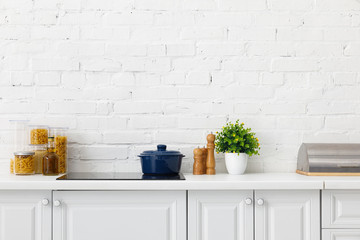 modern white kitchen interior with pot on electric induction cooktop near plant and food containers near brick wall